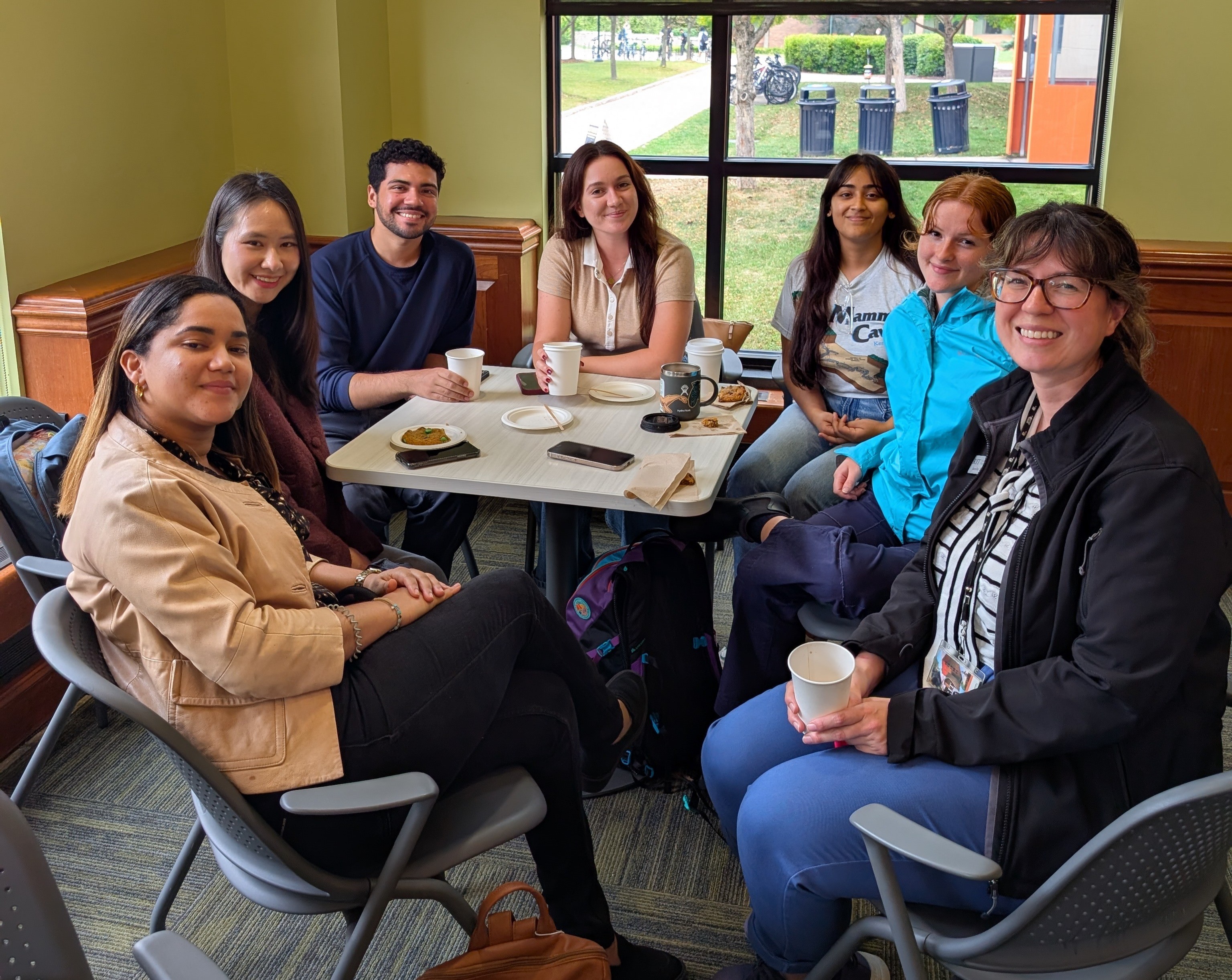 Seven people sit around a table indoors, smiling with drinks and snacks.