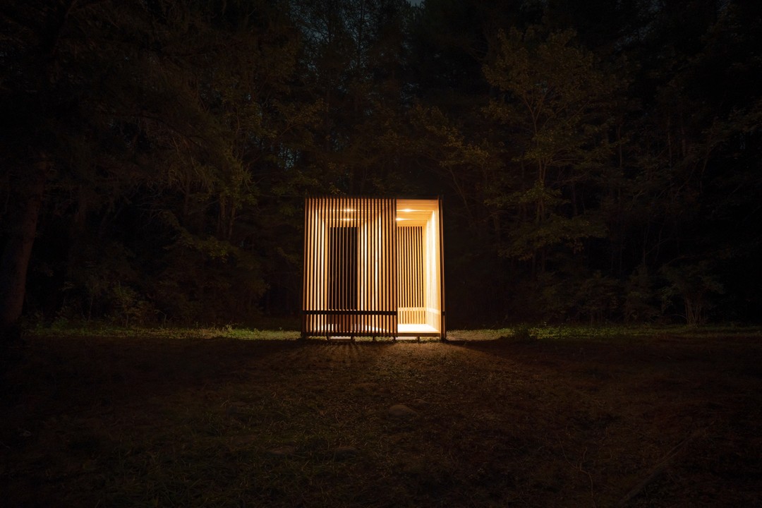 A small, modern wooden pavilion glows warmly in a dark forest, illuminated from within.