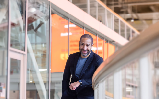Jason Blythe ’02, ’05 MFA smiles while leaning on a railing inside a modern building.