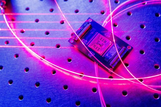 A microchip resting on a perforated optical table, illuminated by vibrant pink and blue light with glowing optical fiber cables looping across the surface.