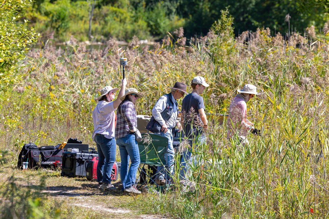 Group of people in hats and casual outdoor clothing conducting field research in tall grass with equipment cases and tools.