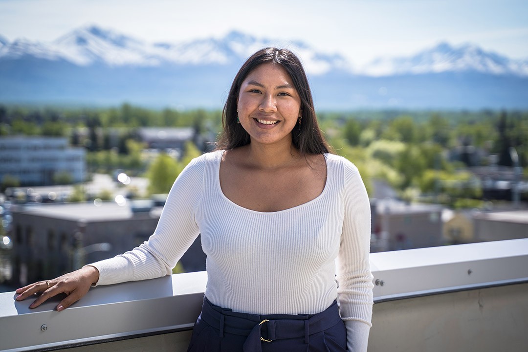 Woman smiling on a balcony with mountains and greenery in the background.