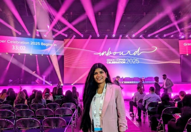 Woman smiling in a pink blazer stands inside a brightly lit auditorium with a large “Grace Hopper Celebration 2025” stage in the background.