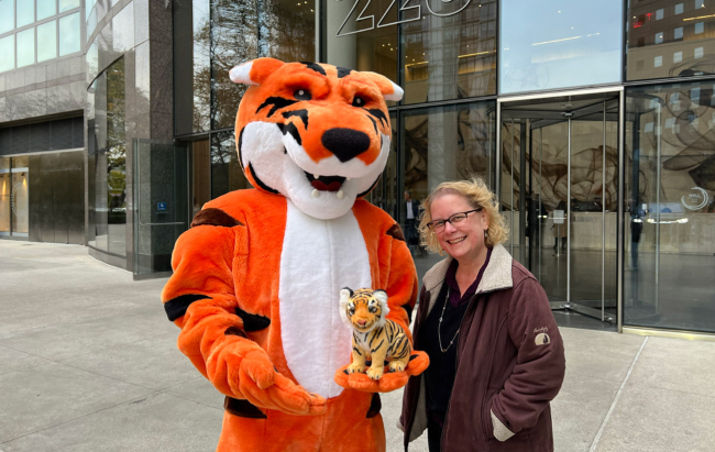 A person in an orange tiger mascot costume stands outside a building beside a smiling woman holding a small stuffed tiger.
