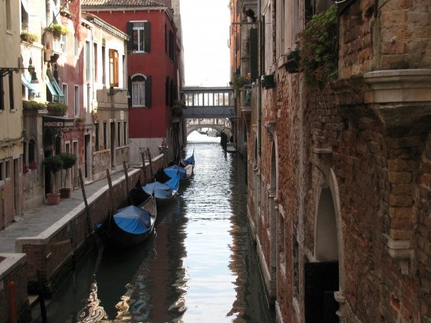 Narrow canal with gondolas beside old buildings and a small bridge ahead.