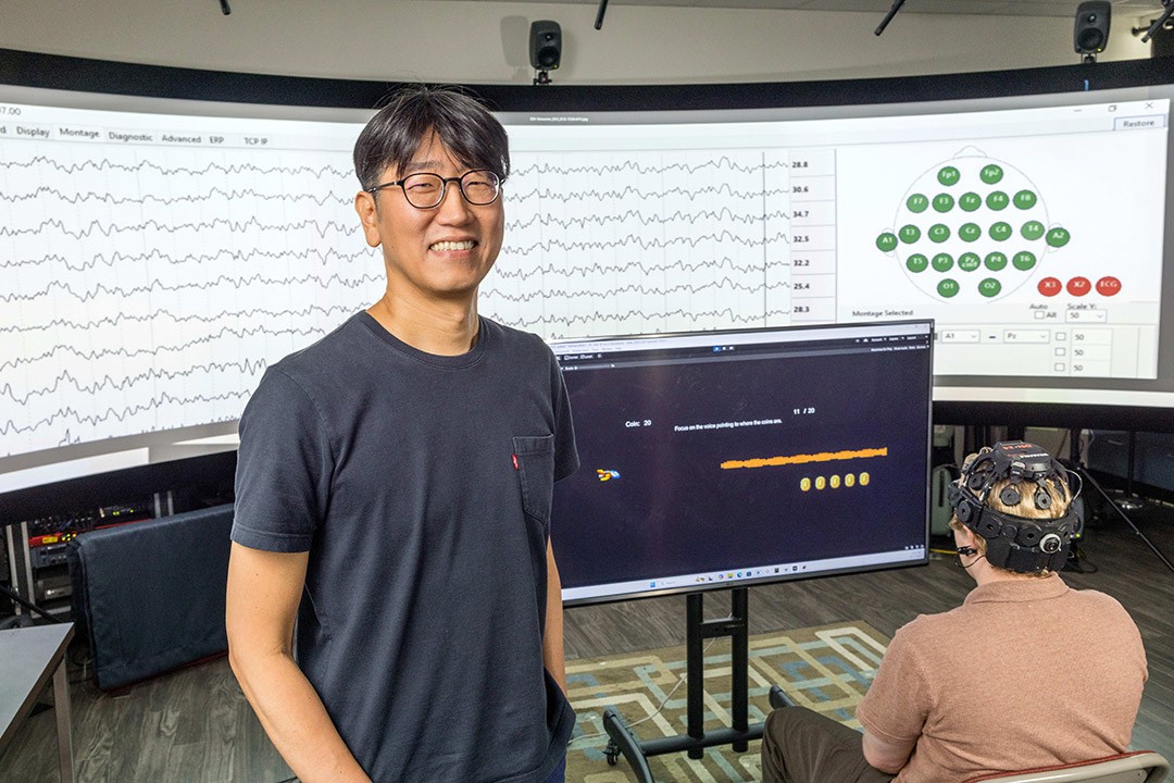 Audio engineer in lab with EEG data displayed and a participant wearing an EEG cap.