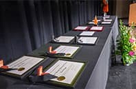 A row of certificates and awards displayed on a black-draped table with flowers in the foreground.