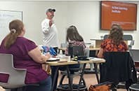 Faculty standing at the front of room presenting with a few faculty members seated
