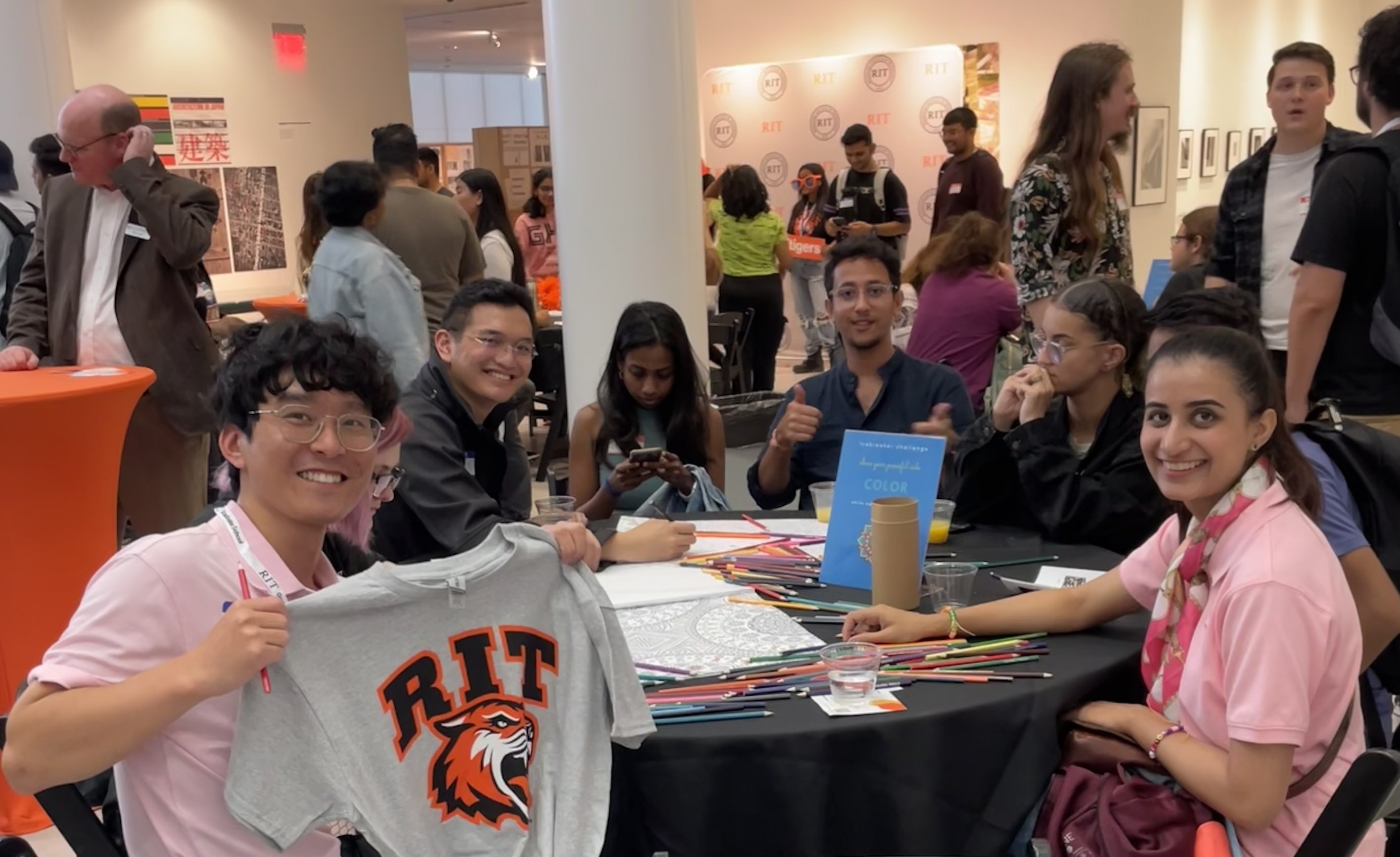 New graduate students sitting on a circular table with one student holding up and showing a gray RIT T-shirt while others smile at the camera