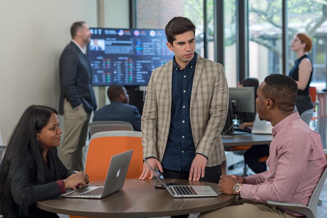 A  woman and two men, engage in a discussion in a modern office setting with digital displays in the background.