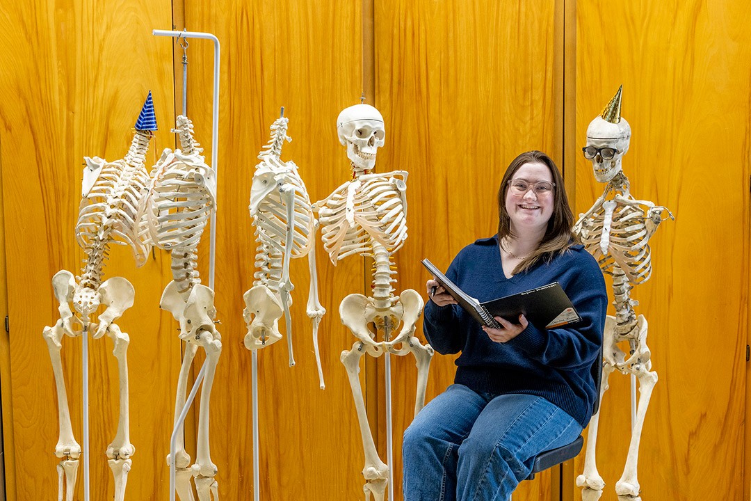 A woman sits among anatomical skeletons, smiling as she holds a notebook.