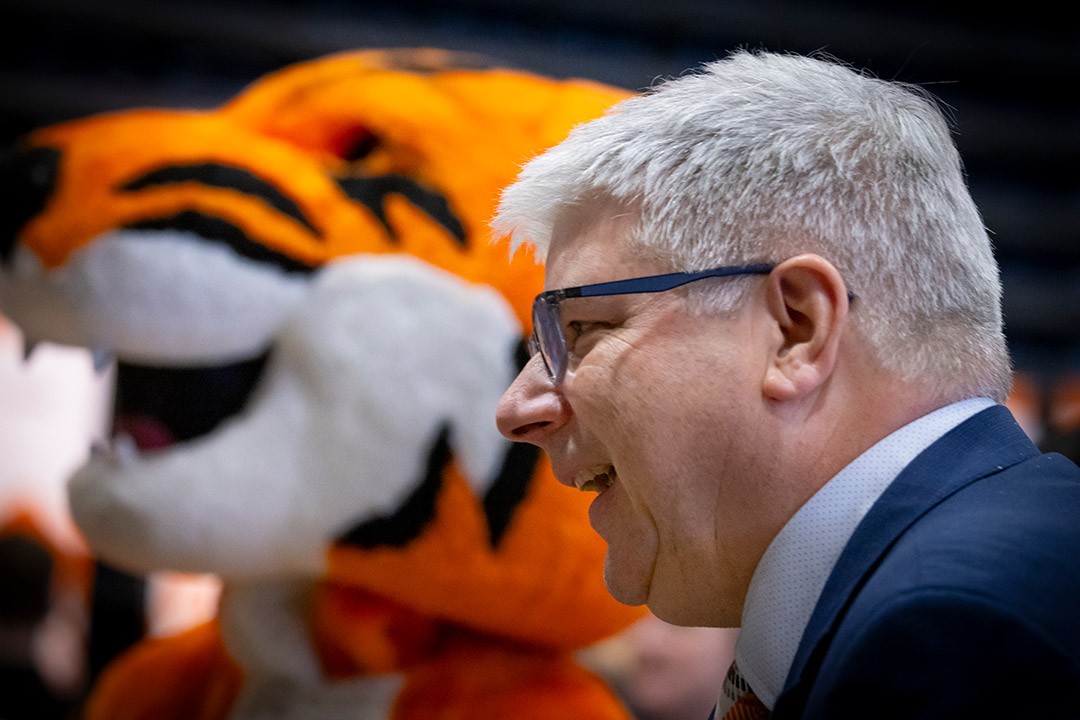 A man in glasses smiling with a tiger mascot in the background.
