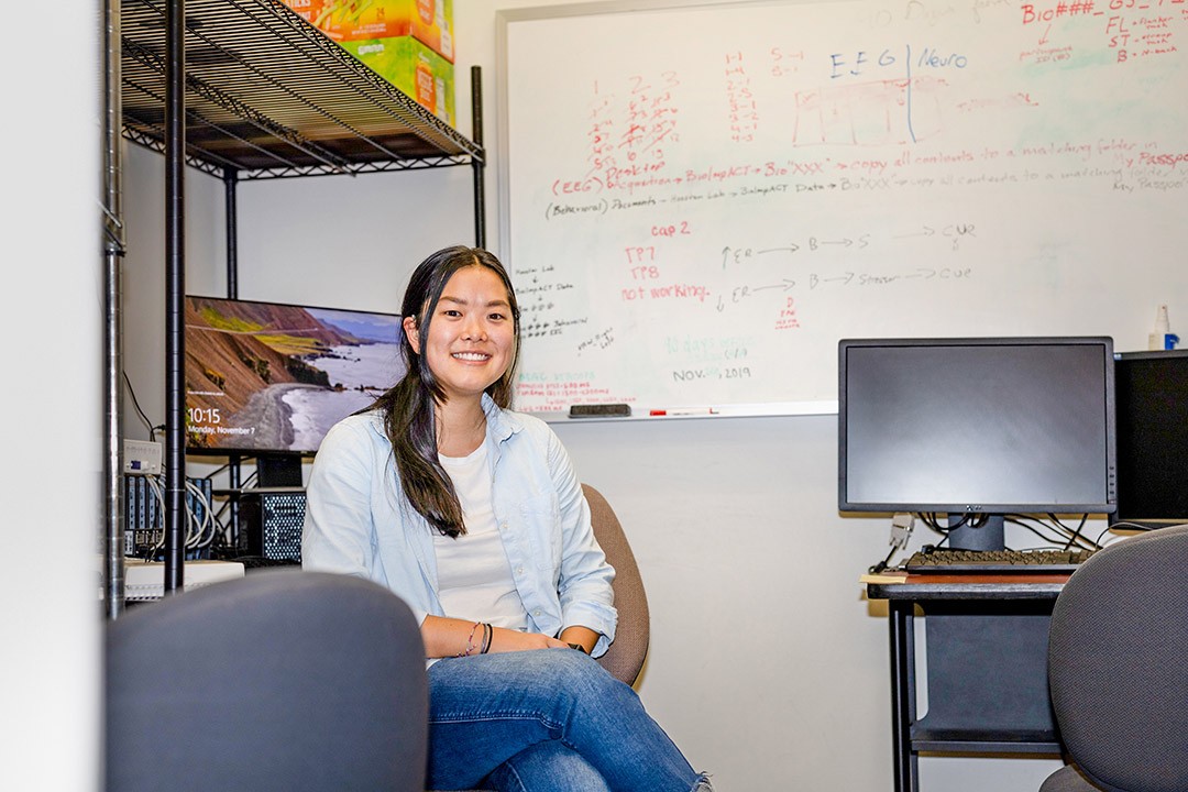 A young woman smiling while seated in a research lab office, with a whiteboard full of notes behind her.
