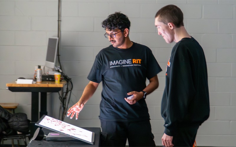 Melson Miranda, wearing an "IMAGINE RIT" t-shirt, gestures toward an illuminated touchscreen display while explaining a project to another person in a black sweatshirt.