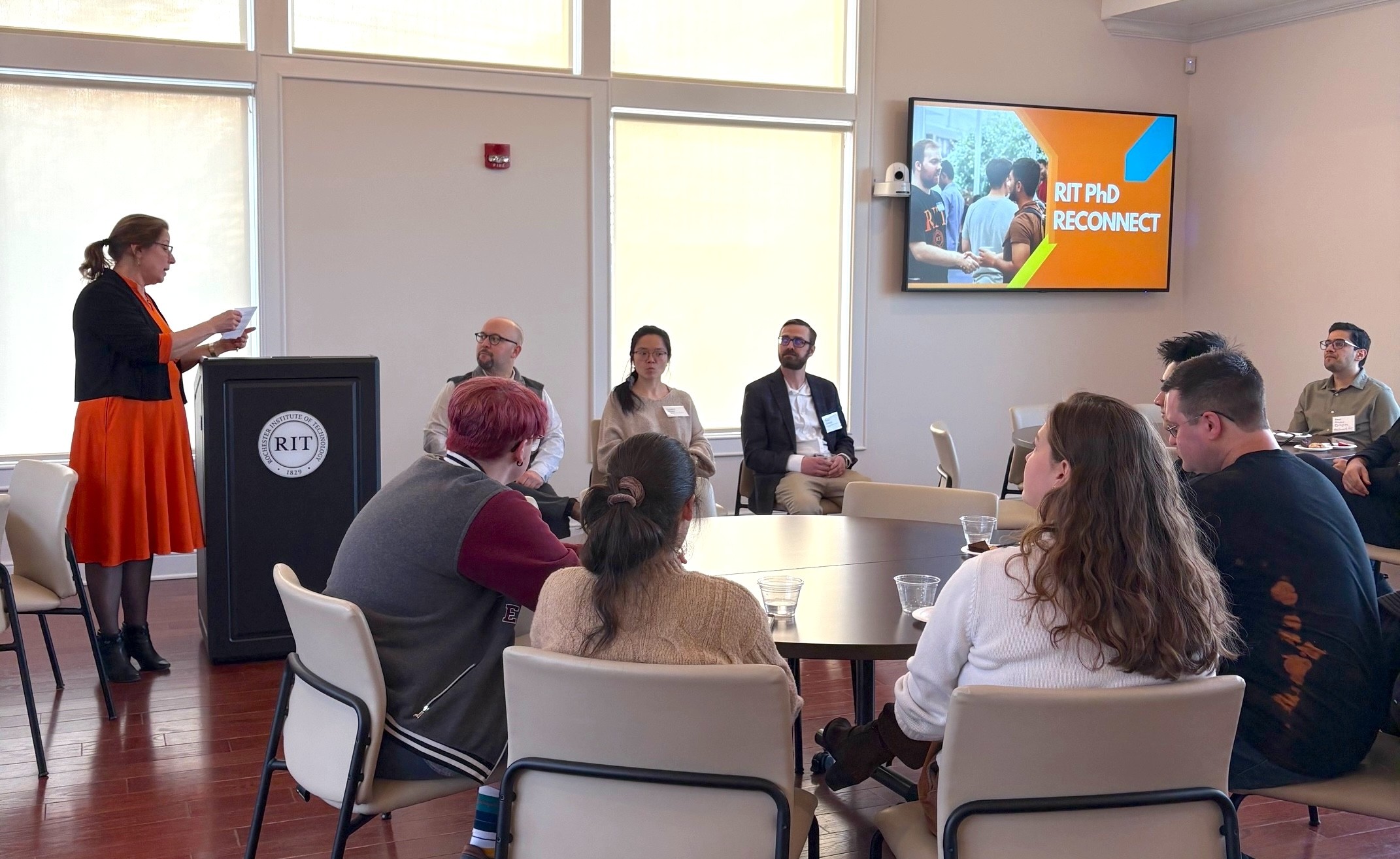 Dean Diane Slusarski addresses attendees at the RIT PhD Reconnect event, with alumni panelists seated beside her and students listening around round tables.