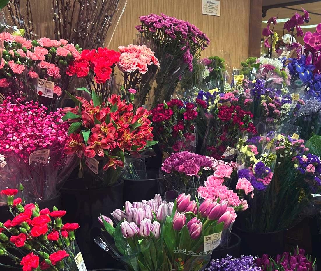 A vibrant display of various fresh-cut flowers in a market, featuring tulips, carnations, and other colorful blossoms wrapped in plastic sleeves.