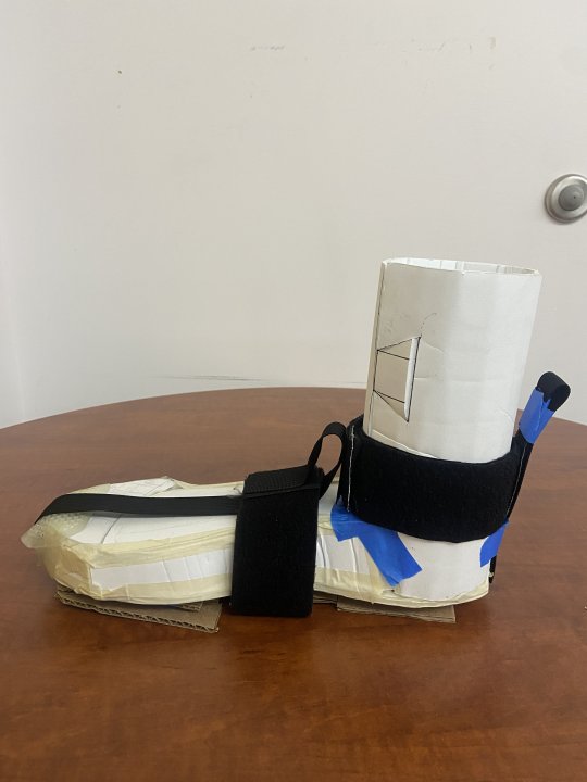 Photo of device prototype: model foot made of posterboard with the cardboard pieces and webbing strap attached. The webbing straps run over the top of the foot from toe to ankle, over the arch of the foot, and around the ankle