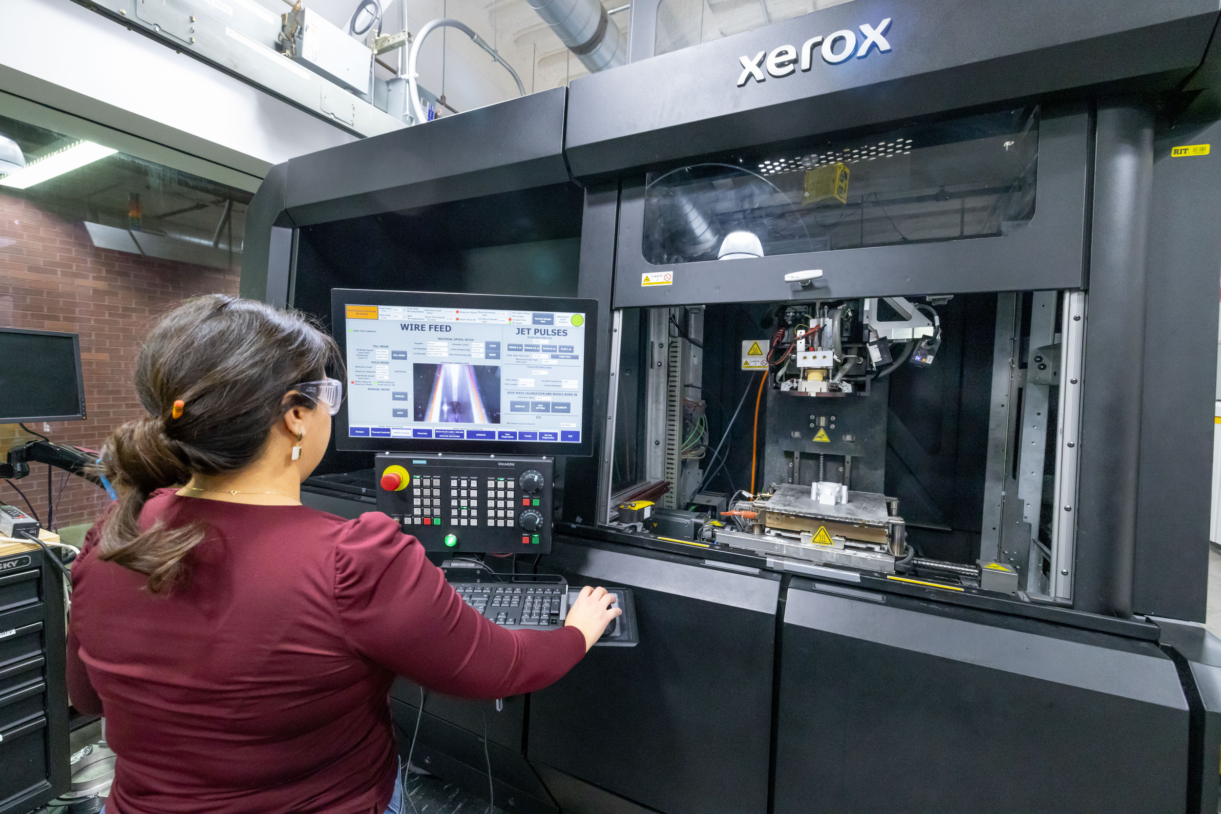 An engineering student standing at a laptop stand attached to a metal 3D printer.