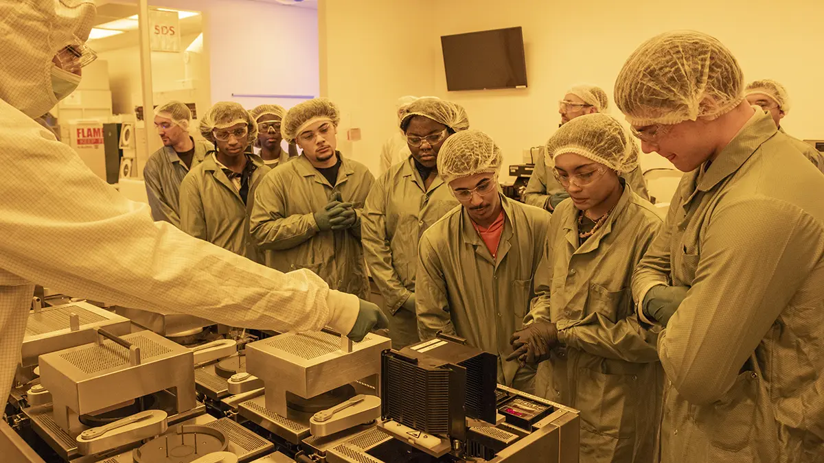 A professor pointing to a piece of equipment in a clean room as high school students observe.
