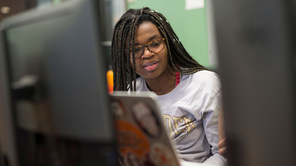 A student wearing glasses and a casual sweatshirt works on a laptop in a computer lab.