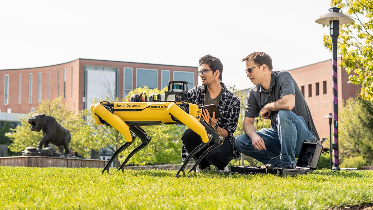 Two researchers operate a Boston Dynamics robotic dog on a grassy field outside a modern campus building.
