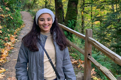 A student wearing a gray jacket and headband, outside in the woods, standing in front of a tree and a wooden fence.