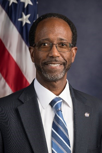 A headshot of Clayton Turner wearing a navy suit with an United States flag in the background.