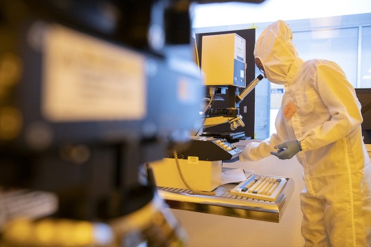 A person wearing a clean room suit and looking into a microscope.