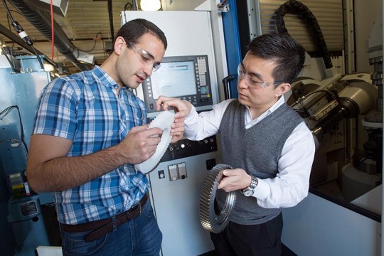 A student in a plaid shirt holding a plastic gear and a faculty member holding a metal gear.