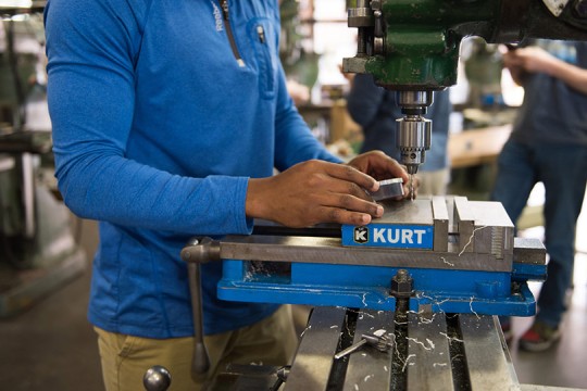 Student wearing a blue shirt working with a drill press.