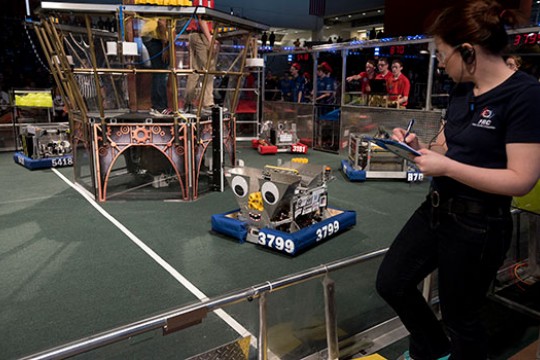 A person writing on a clipboard while looking at a robot.