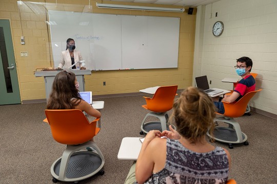 professor wearing a mask stands in the front of a classroom behind a plexiglas barrier as three students sit six feet apart.