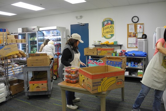 A person standing at a table in a food cupboard room with fruit and food stuffs.
