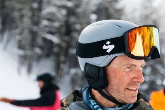 a man stands near a ski slope with a gray ski helmet and black goggles on his head.