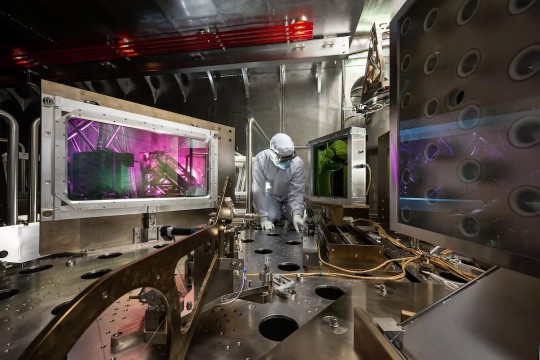 Technician in cleanroom suit adjusts equipment inside high-tech laboratory chamber with glowing lasers.