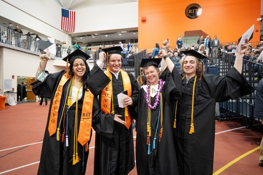 Four college graduates in an arena smile and cheer while wearing black caps and gowns.