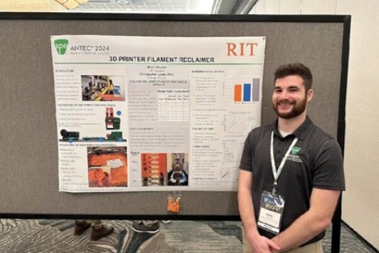 a young man in a black shirt stands in front of a mastrs capstone poster in a conference room