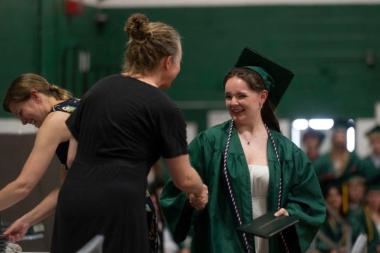 Woman in green college graduation robes receives a diploma and shakes hands with someone.