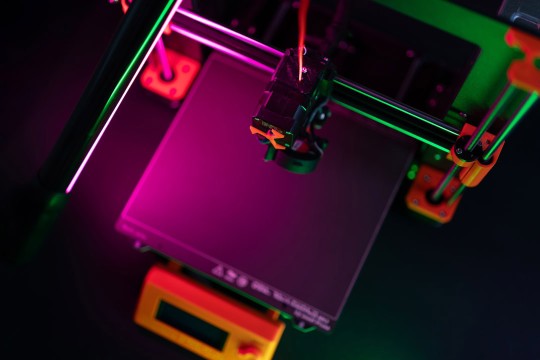 3D printer extruder hovers over empty print bed under neon pink and green lighting.
