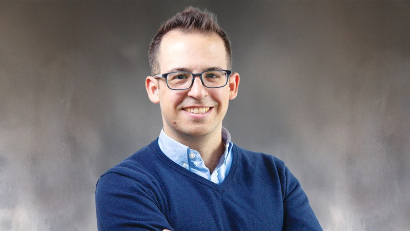 Ryan Larcome is wearing a dark blue sweater over a light blue collared shirt stands with arms crossed against a neutral gray studio background.