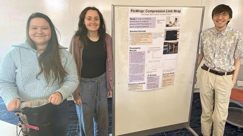 Three students standing on either side of a research poster on FloWrap and smiling.