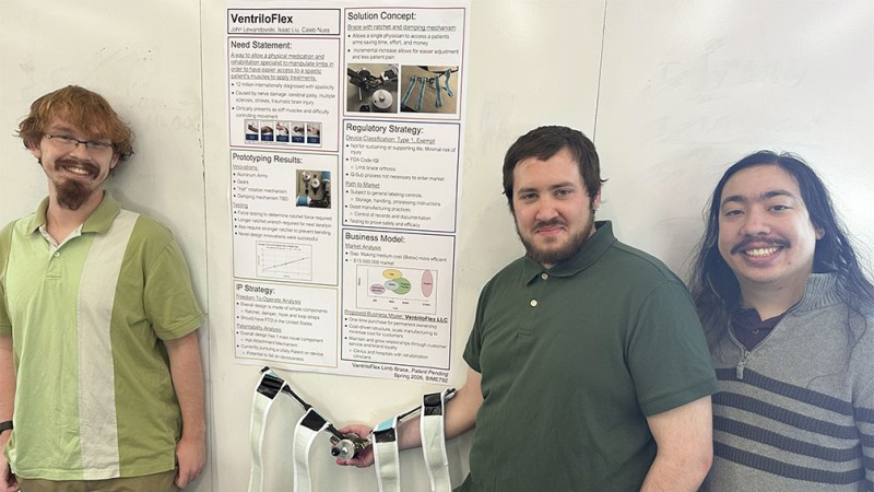 Three students standing on either side of a research poster for VentriloFlex and smiling.