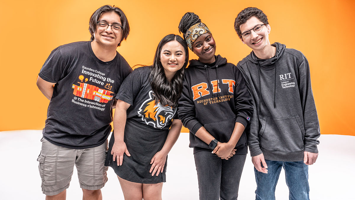 A diverse group of students wearing R I T apparel pose against an orange studio backdrop.