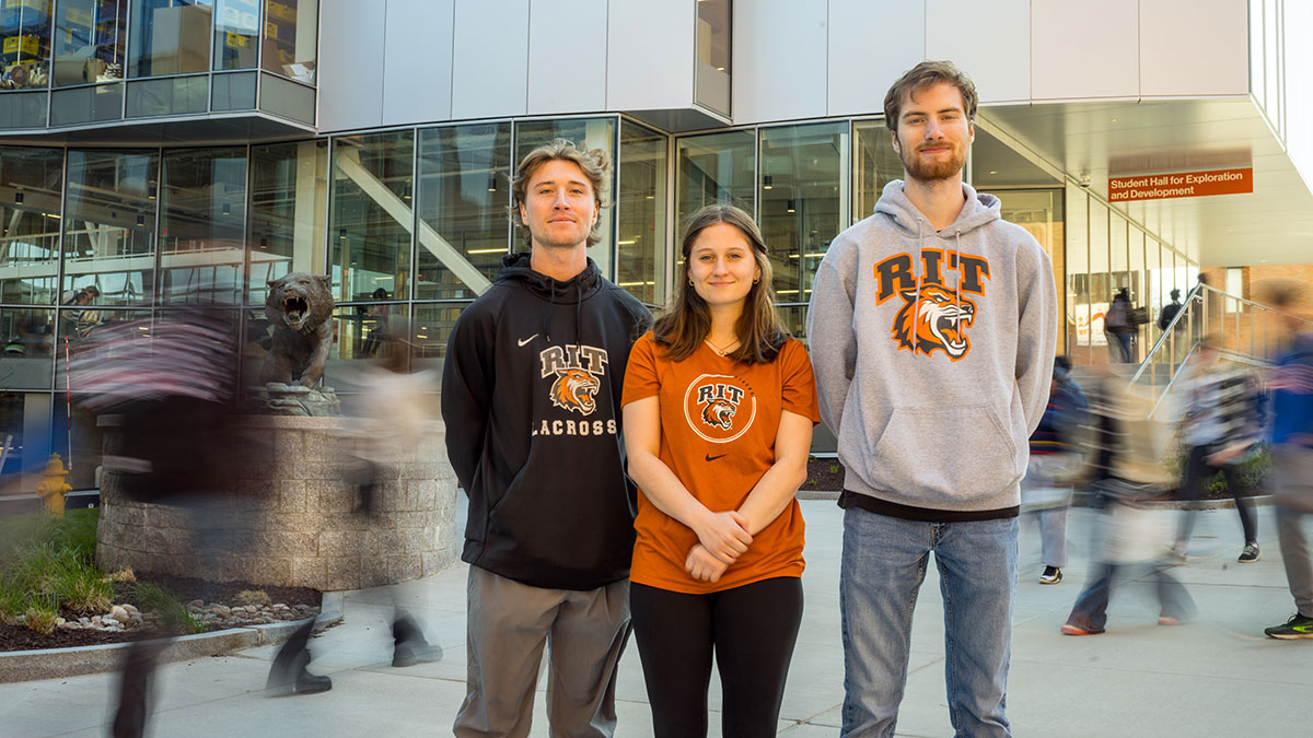 Three students wearing RIT gear stand outside a modern campus building with blurred pedestrian motion in the background.