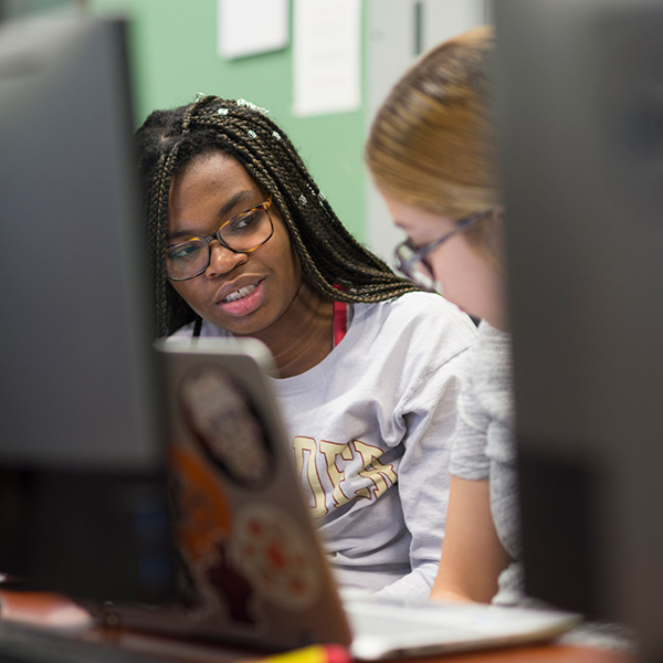 2 students in a computer lab, speaking to eachother.