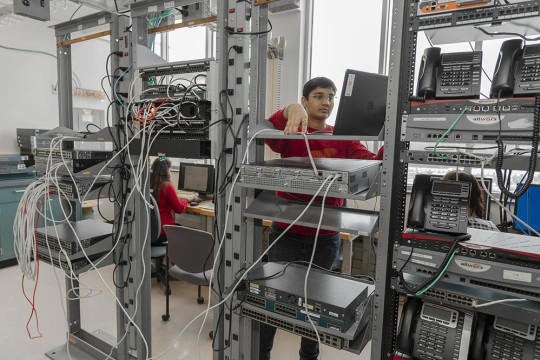 a student working in a server room, with several racks holding communications equipment.