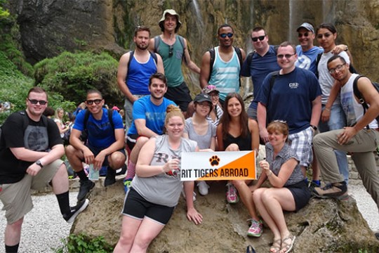 A group of students who traveled abroad pose for a photo on a large boulder holding up a poster that reads "RIT Tigers Abroad."
