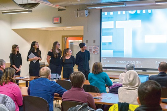 The Jet-2k team stands at the front of a room and shows students and potential "investors" a video about their packaging design.