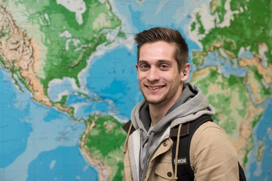 Connor Draughn poses for a photo and smiles in front of a large world map.