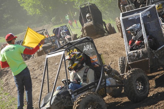 Volunteer waves yellow flag as baja racecars descend a hill.